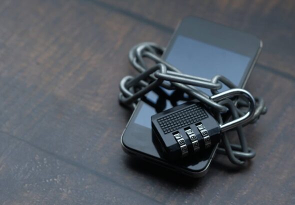 Smartfon owinięty metalowym łańcuchem zapiętym na czarną kłódkę szyfrową na drewnianym stole.