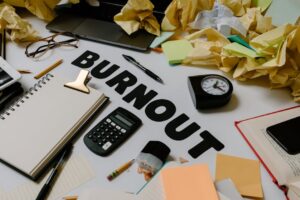 Biurko z napisem „Burnout”, kalkulatorem, zegarem i wieloma pogniecionymi kartkami papieru.