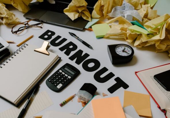 Biurko z napisem „Burnout”, kalkulatorem, zegarem i wieloma pogniecionymi kartkami papieru.