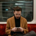 Mężczyzna w brązowym płaszczu siedzi w pociągu i korzysta ze smartfona.