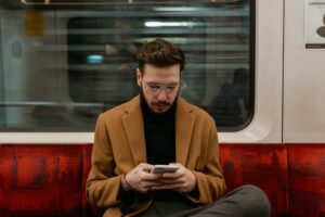 Mężczyzna w brązowym płaszczu siedzi w pociągu i korzysta ze smartfona.