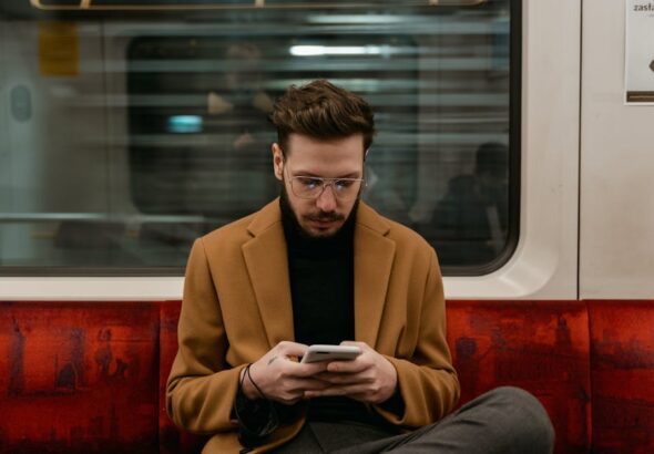 Mężczyzna w brązowym płaszczu siedzi w pociągu i korzysta ze smartfona.
