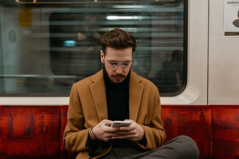 Mężczyzna w brązowym płaszczu siedzi w pociągu i korzysta ze smartfona.