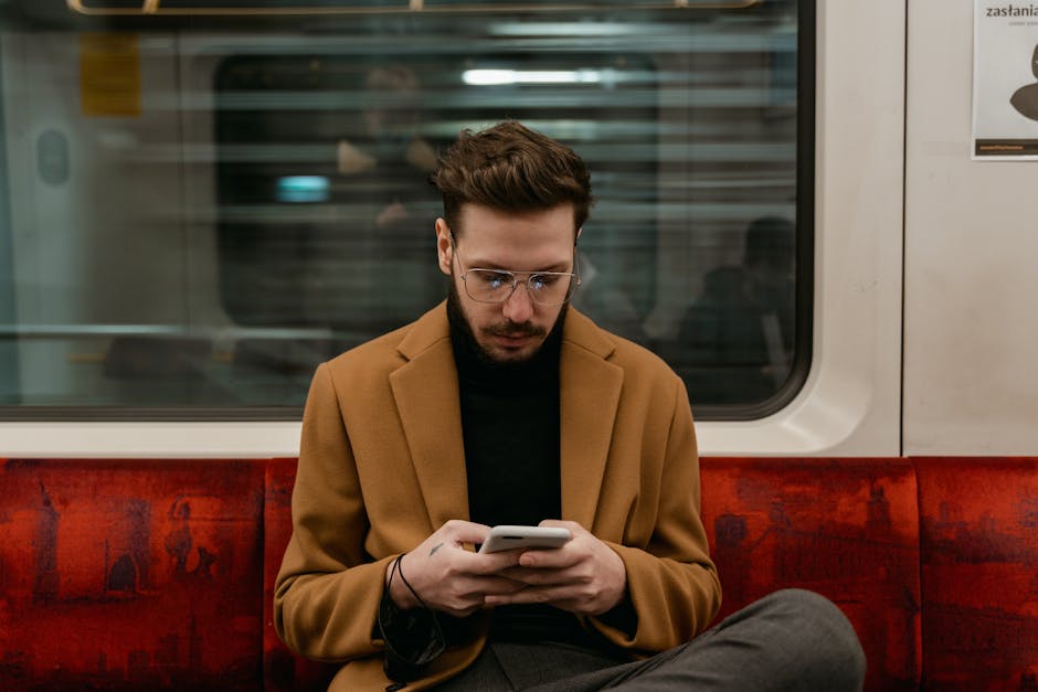 Mężczyzna w brązowym płaszczu siedzi w pociągu i korzysta ze smartfona.