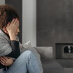 Kobieta z podwiniętymi nogami siedzi na szarej kanapie, trzymając dłoń przy głowie.