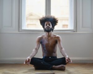 Mężczyzna z afro siedzący w pozycji lotosu na podłodze przed oknem.