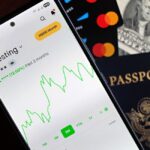 Smartfon z wykresem giełdowym, paszport oraz karty płatnicze na blacie.