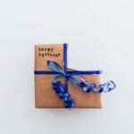 Prezent w brązowym papierze owinięty niebieską wstążką z napisem „happy birthday”.
