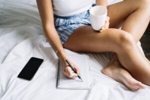 Kobieta siedząca na łóżku pisze w notesie, trzymając kubek z napojem.